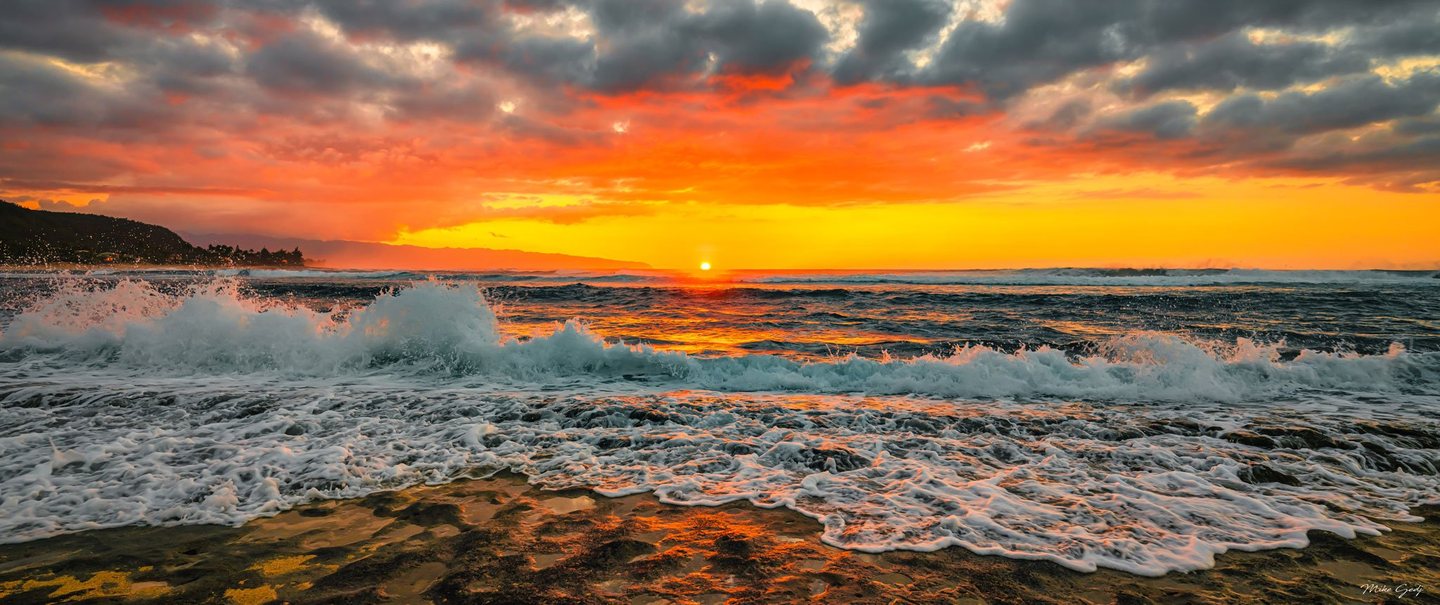 Waves break across a rocky shoreline beneath an orange and gold sky, with the sun low on the ocean horizon and a dark headland to the left.
