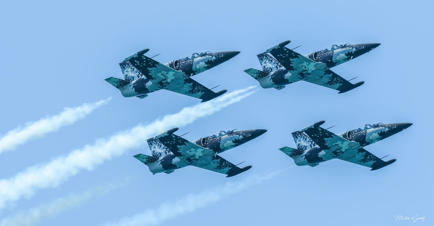 Four jets with dark blue, turquoise, and white patterned markings fly in close formation across a pale blue sky, trailing white smoke.