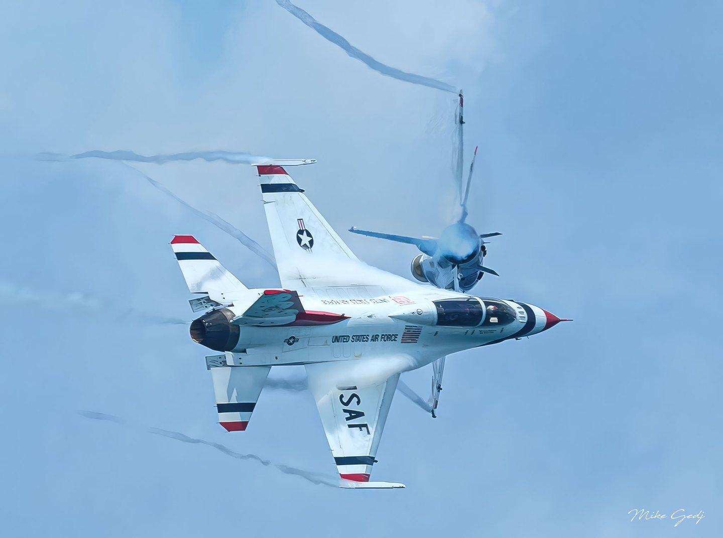 Two U.S. Air Force fighter jets bank against a pale blue sky, with a white, red, and blue aircraft dominating the foreground and vapor curling around the jet behind it.