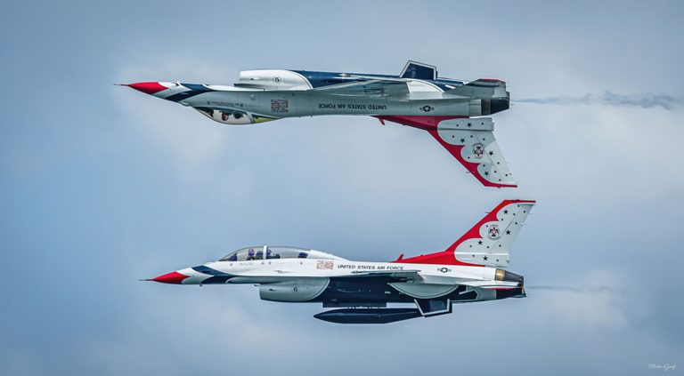 Two red, white, and blue United States Air Force Thunderbird jets fly leftward in parallel, with the upper aircraft inverted above the lower against a pale blue sky.