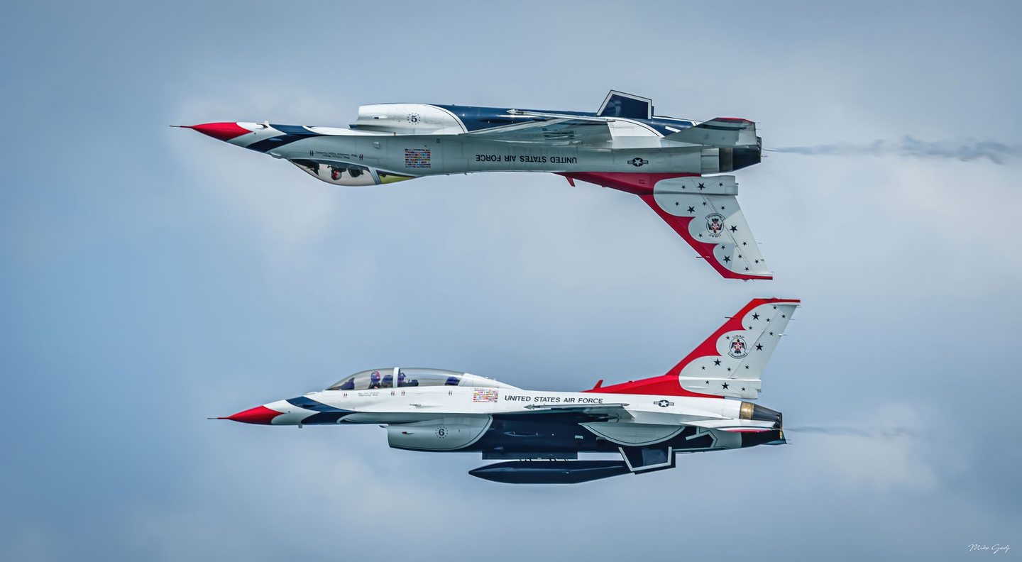 Two red, white, and blue United States Air Force Thunderbird jets fly leftward in parallel, with the upper aircraft inverted above the lower against a pale blue sky.