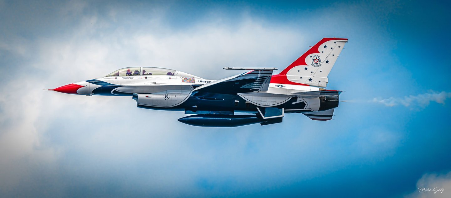 A U.S. Air Force Thunderbirds F-16 jet with red, white, and dark blue markings flies nose-left against a blue sky and soft white clouds.