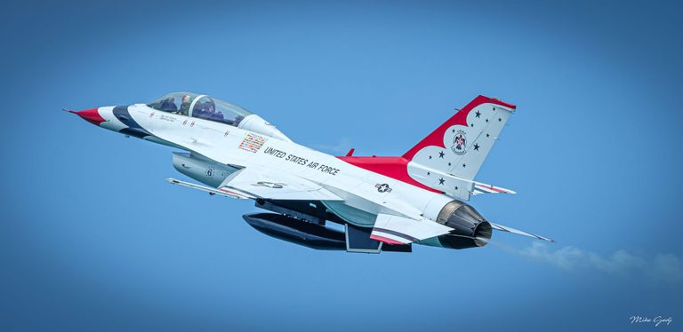 A white, red, and blue U.S. Air Force Thunderbirds F-16 jet banks against a clear blue sky, its nose pointing toward the upper left.