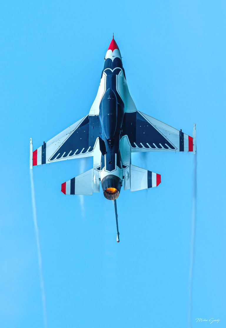 A red, white, and dark blue fighter jet seen from below points upward against a clear blue sky, with an orange exhaust glow and faint trails behind its wings.