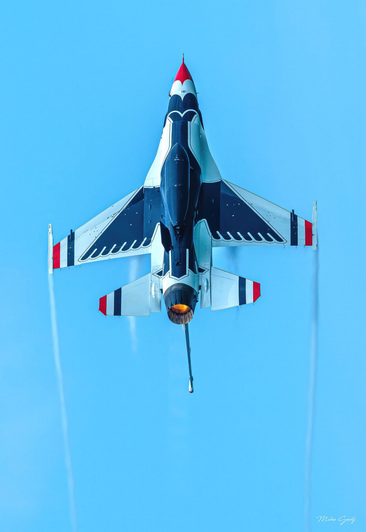 A red, white, and dark blue fighter jet seen from below points upward against a clear blue sky, with an orange exhaust glow and faint trails behind its wings.