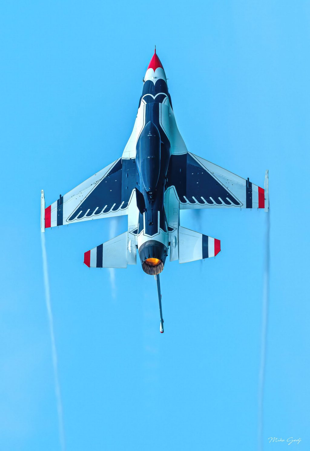 A red, white, and dark blue fighter jet seen from below points upward against a clear blue sky, with an orange exhaust glow and faint trails behind its wings.