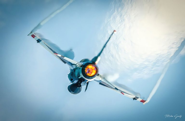 A fighter jet seen from behind banks across a pale blue sky, its orange afterburner glowing between swept wings trailing white vapor.