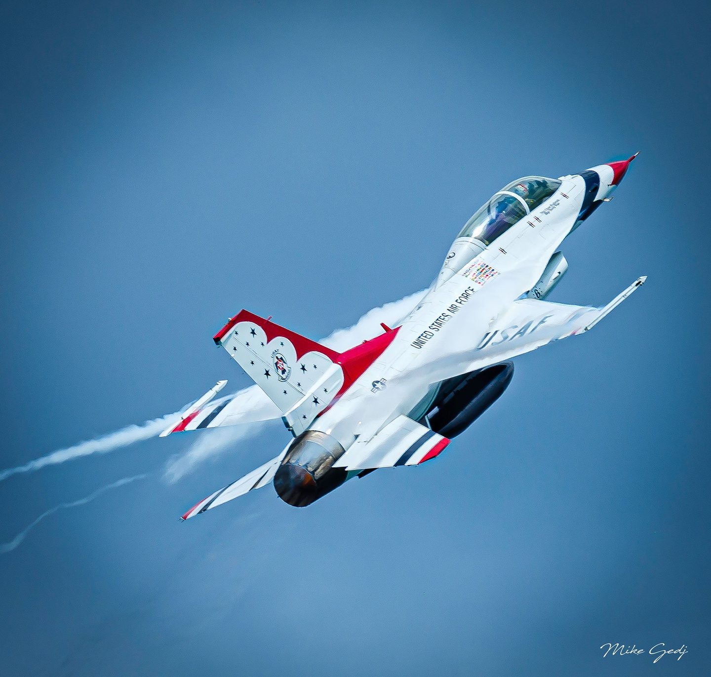 A white, red, and blue United States Air Force Thunderbirds jet banks against a clear blue sky with white vapor streaming behind its wings.