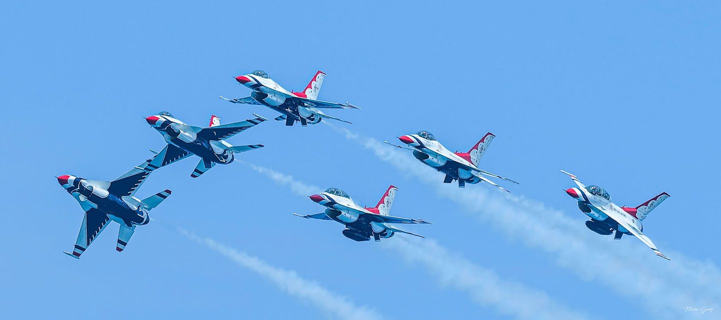Six red, white, and blue fighter jets bank across a clear blue sky, leaving pale smoke trails behind them.