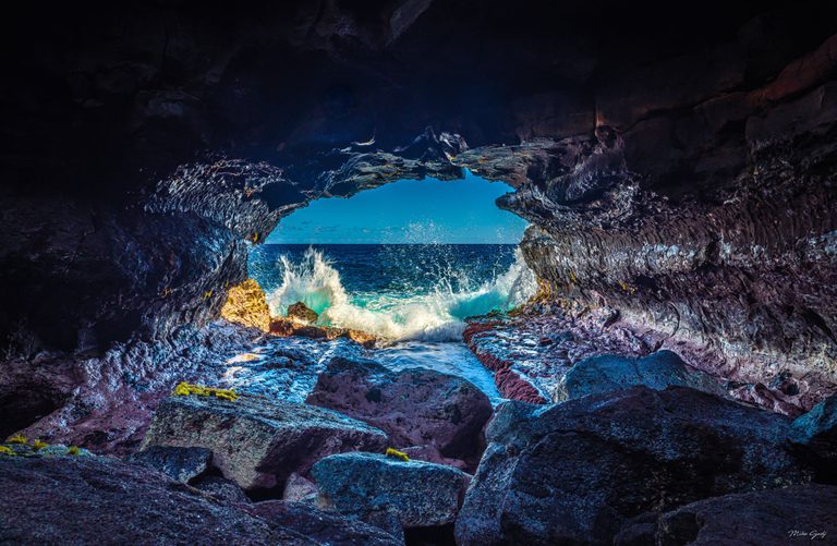 Waves break against rocks at the mouth of a dark sea cave, framing a blue ocean horizon and bright sky beyond.