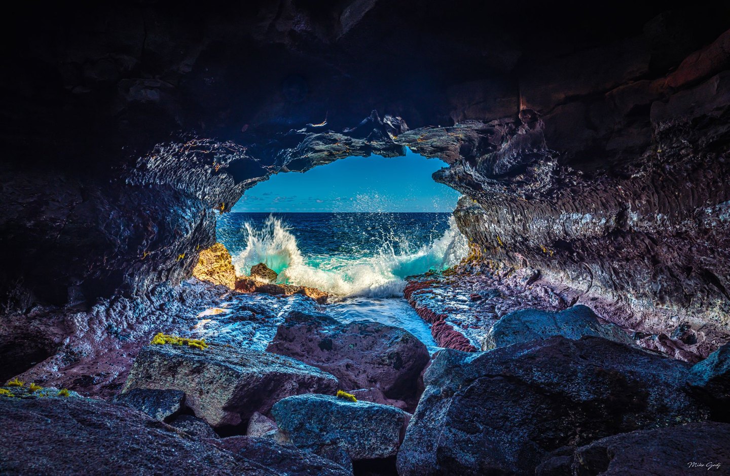 Waves break against rocks at the mouth of a dark sea cave, framing a blue ocean horizon and bright sky beyond.
