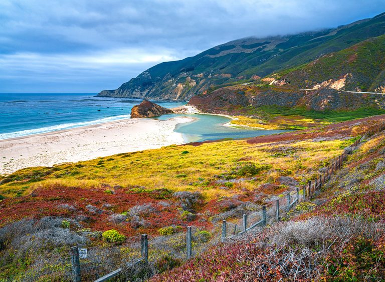 A turquoise coastal lagoon curves behind a pale sandy beach beneath cloud-covered mountains, with colorful vegetation and a wire fence in the foreground.