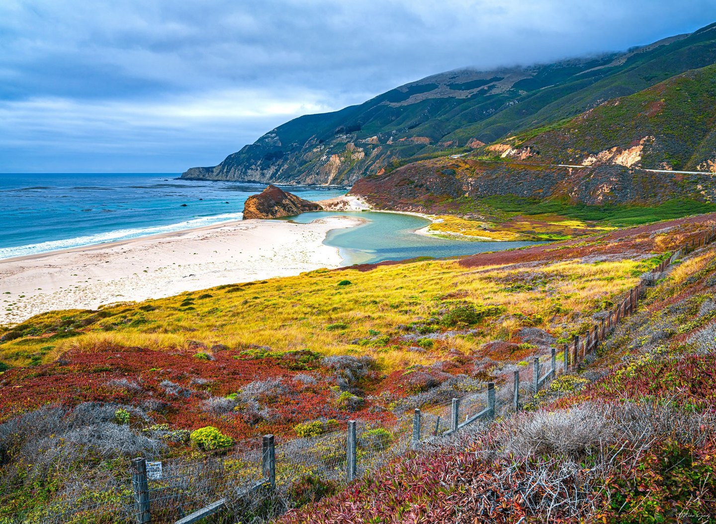A turquoise coastal lagoon curves behind a pale sandy beach beneath cloud-covered mountains, with colorful vegetation and a wire fence in the foreground.