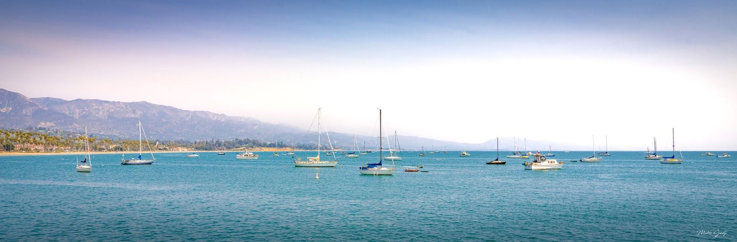 Sailboats and small motorboats rest on turquoise coastal water beneath a pale blue sky, with a palm-lined shore and hazy mountains in the distance.