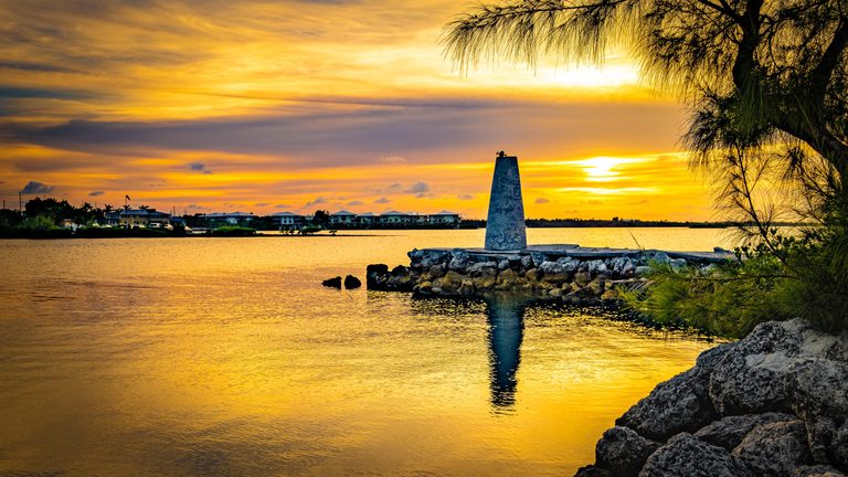 A tapered concrete pillar on a rocky waterfront reflects in golden water beneath a sunset sky, with distant houses and overhanging tree branches.