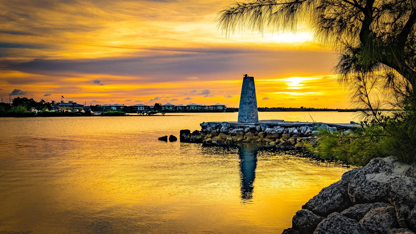 A tapered concrete pillar on a rocky waterfront reflects in golden water beneath a sunset sky, with distant houses and overhanging tree branches.