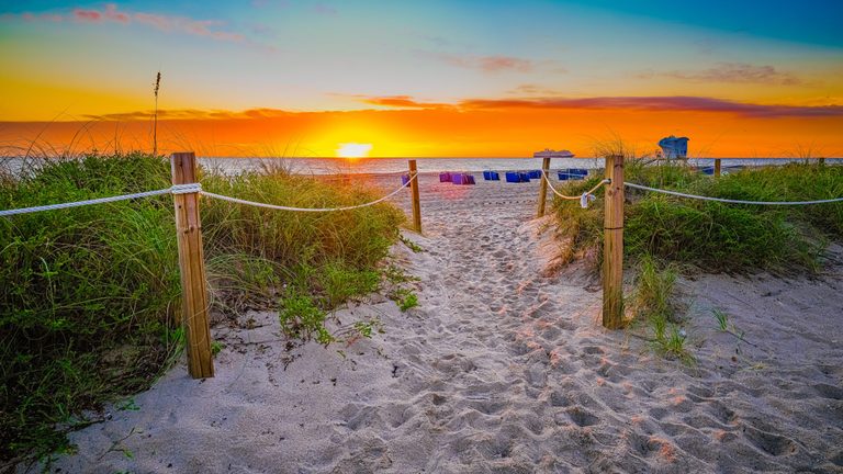 A sandy path bordered by rope fences and dune grasses leads to an ocean beach beneath an orange sky, with a low sun, blue lifeguard tower, and distant cruise ship.