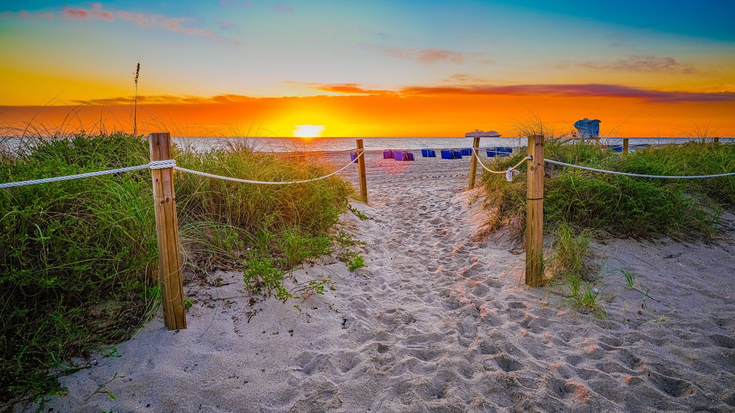 A sandy path bordered by rope fences and dune grasses leads to an ocean beach beneath an orange sky, with a low sun, blue lifeguard tower, and distant cruise ship.