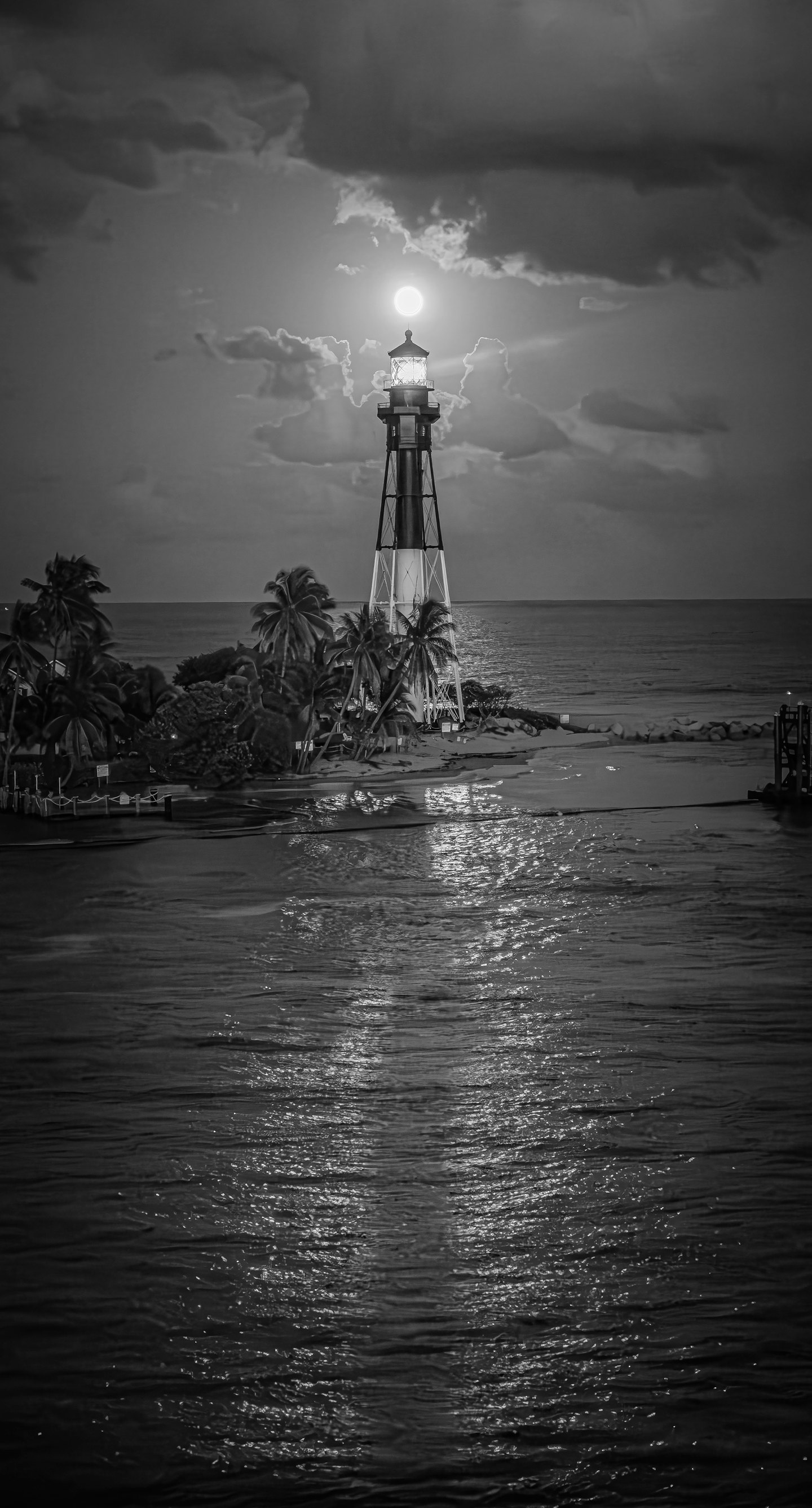 Black-and-white view of a lighthouse beneath a bright sun, with palms, clouds and shimmering coastal water.