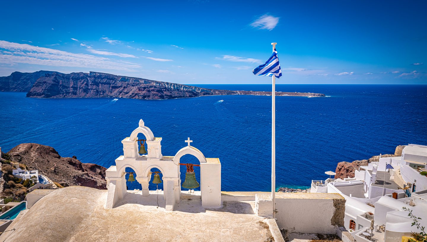 Whitewashed arches hold four bells above a deep blue sea, with a Greek flag and rugged cliffs beyond.