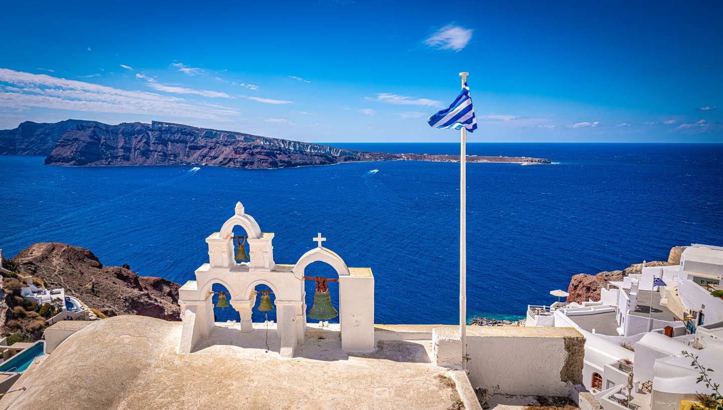 Whitewashed arches hold four bells above a deep blue sea, with a Greek flag and rugged cliffs beyond.