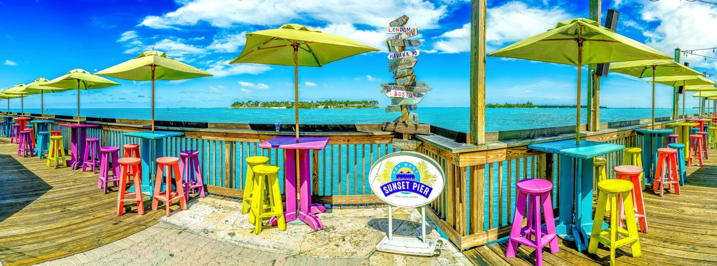 Lime umbrellas shade colorful tables and stools beside turquoise water at Sunset Pier in Key West.
