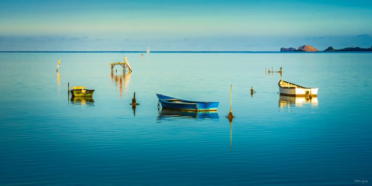 Three small boats reflect in calm turquoise water, with mooring posts, a raised platform and distant rocky islands.