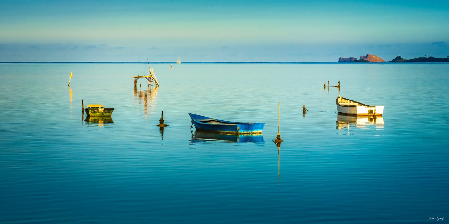 Three small boats reflect in calm turquoise water, with mooring posts, a raised platform and distant rocky islands.