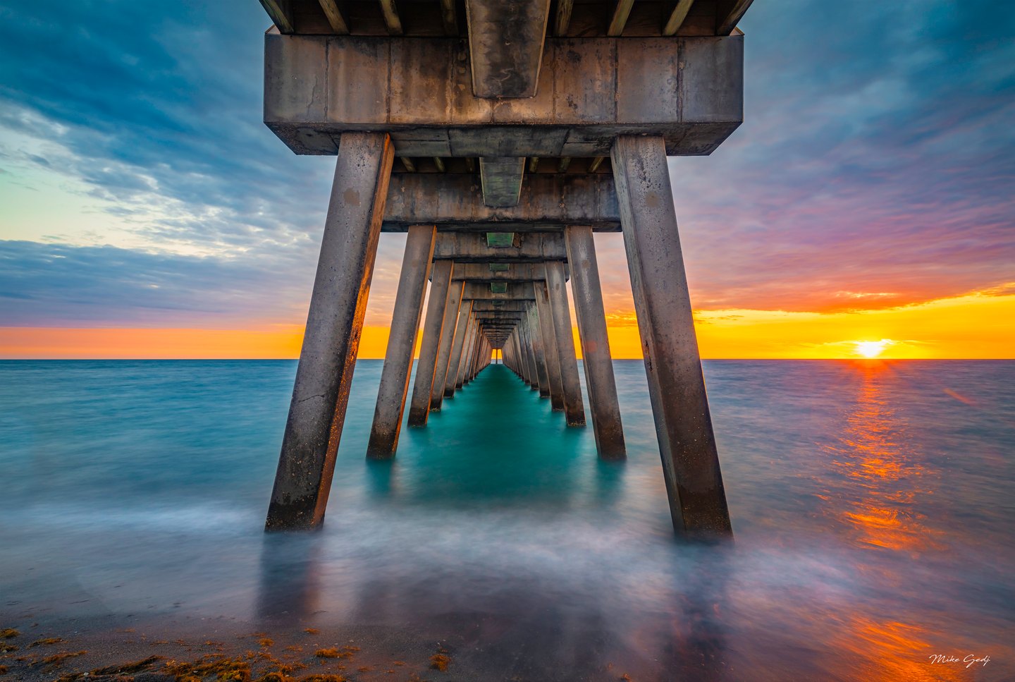 Concrete pier supports recede over turquoise water beside a low sun, with orange reflections across the wet shore.