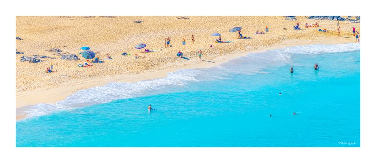 An elevated view of swimmers in turquoise surf beside a sandy beach dotted with colorful umbrellas and rocks.