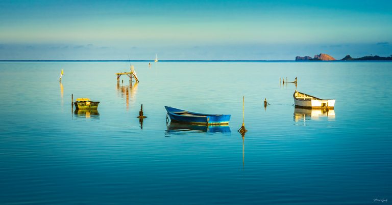 Three small boats reflect in calm turquoise water, with wooden posts, a raised platform and distant rocky land.