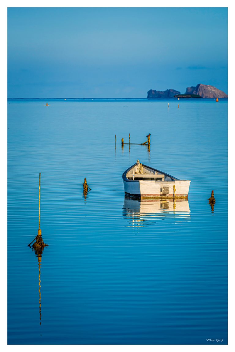 A white boat rests among wooden posts in calm turquoise water, with reflections, buoys, and distant rocky islands.