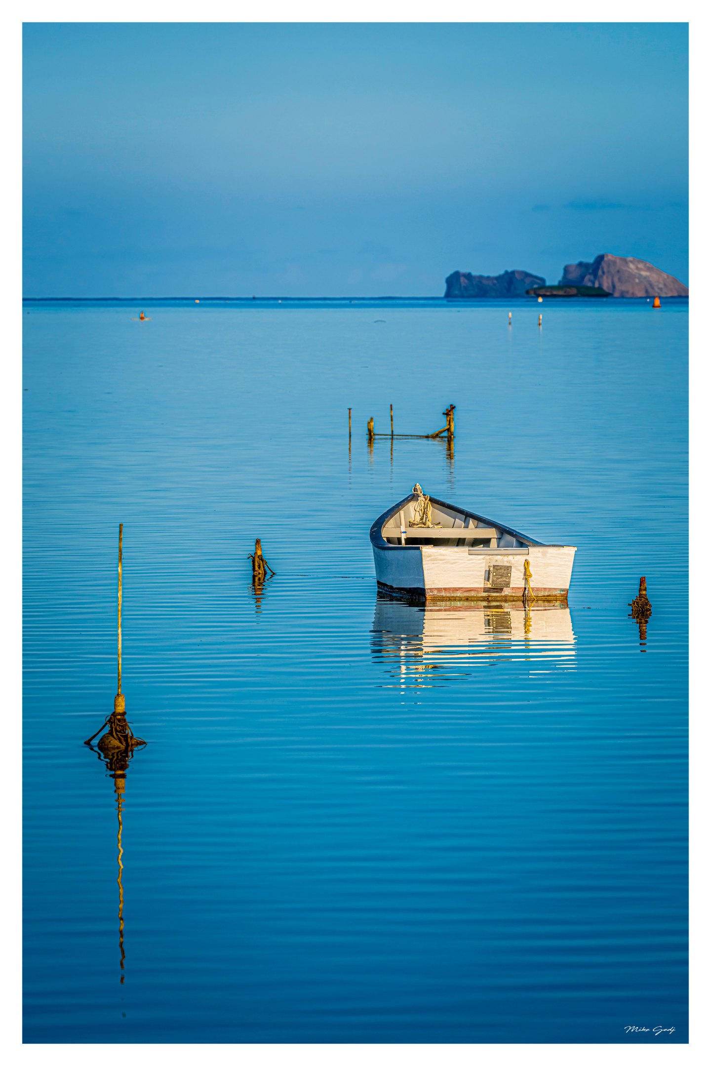 A white boat rests among wooden posts in calm turquoise water, with reflections, buoys, and distant rocky islands.