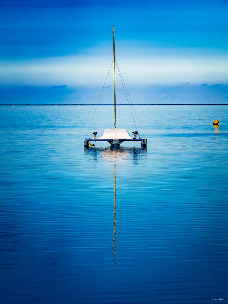 A white multihull sailboat rests on blue water, its bare mast reflected below, with a yellow buoy to the right.