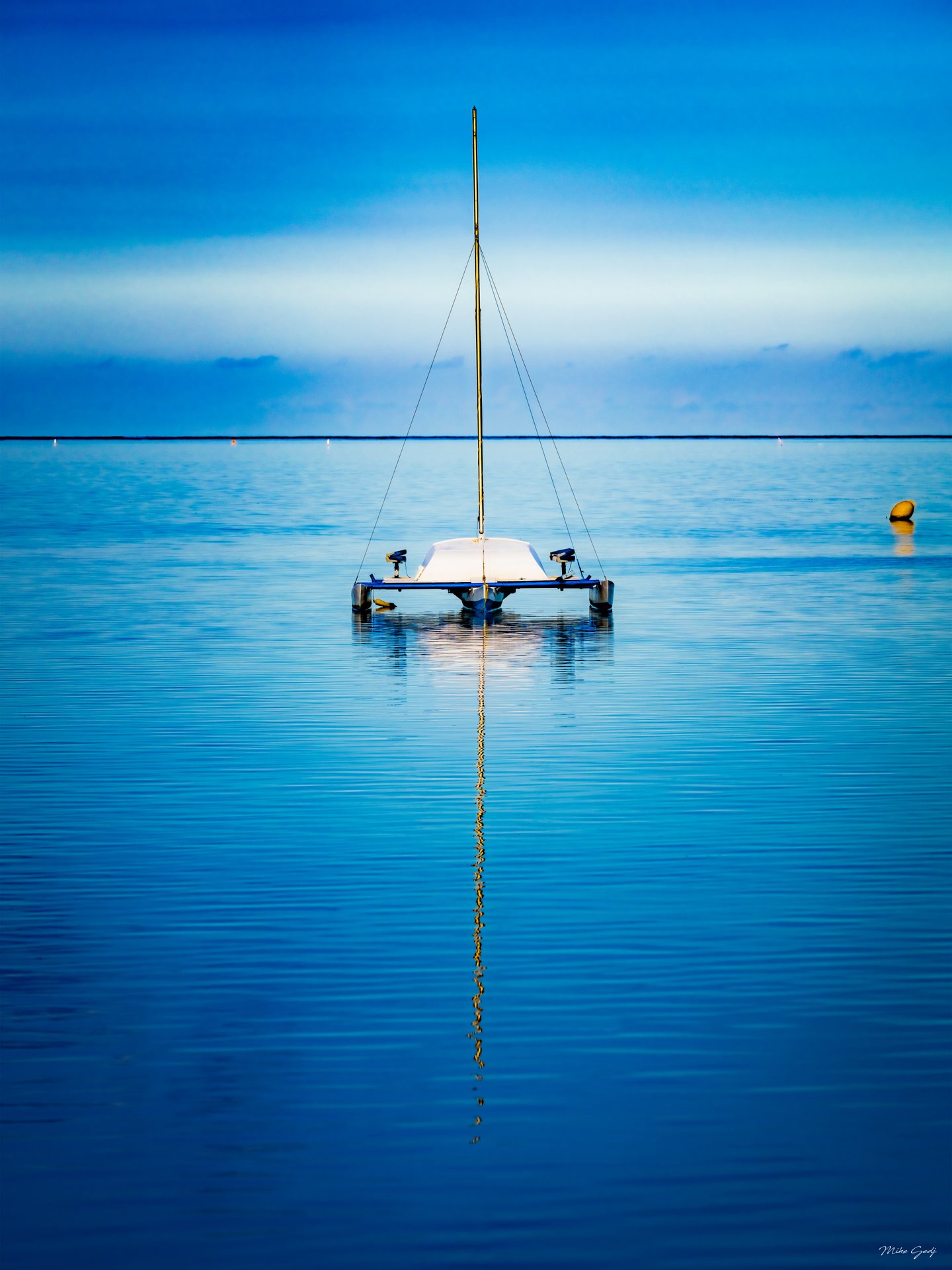 A white multihull sailboat rests on blue water, its bare mast reflected below, with a yellow buoy to the right.