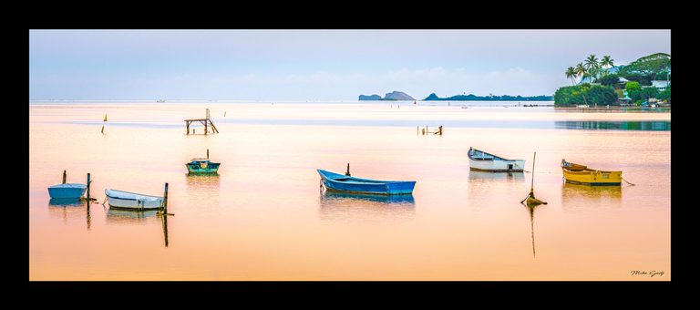 Colorful boats moored in peach-toned water, with a laddered platform, distant rocky islets, and a palm-lined shore.