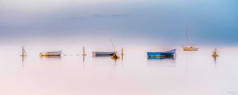 Three small boats and a sailboat sit among mooring posts, reflected in calm water beneath a pale blue sky.