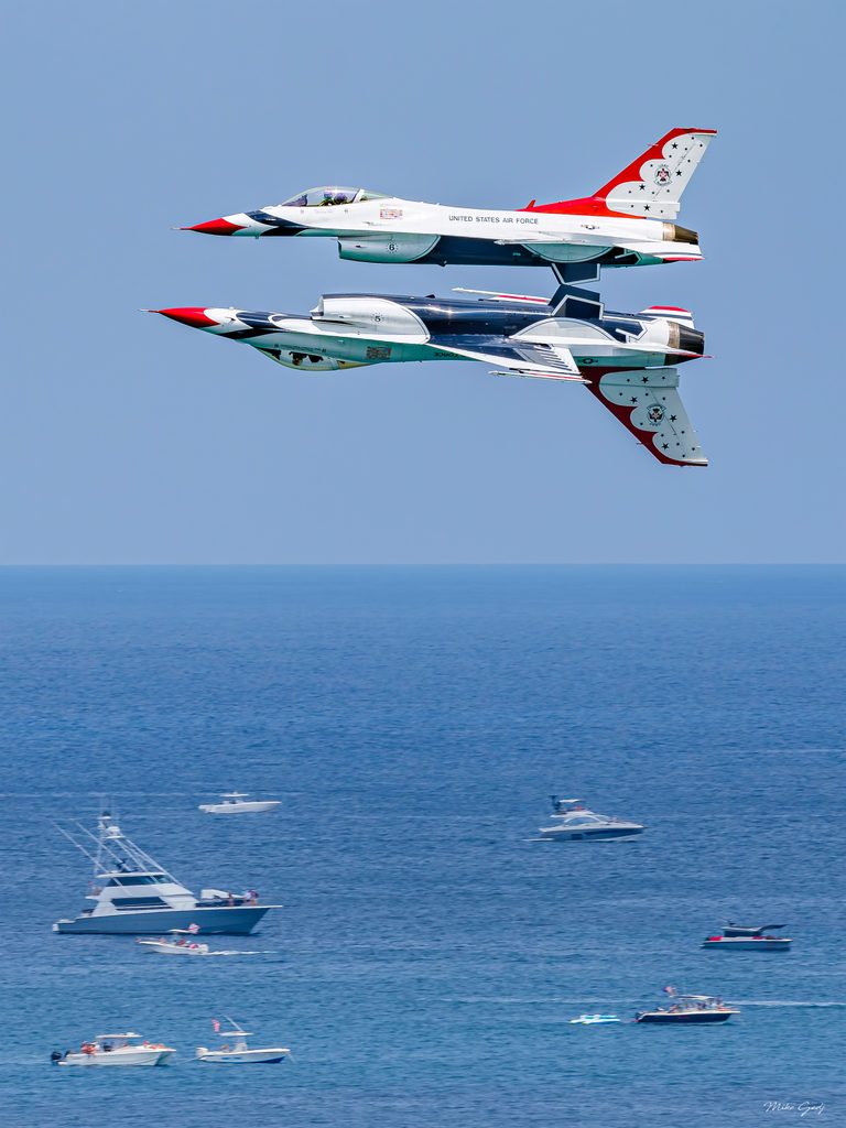 Two red, white and blue jets fly close together, one inverted, above a blue sea dotted with motorboats.