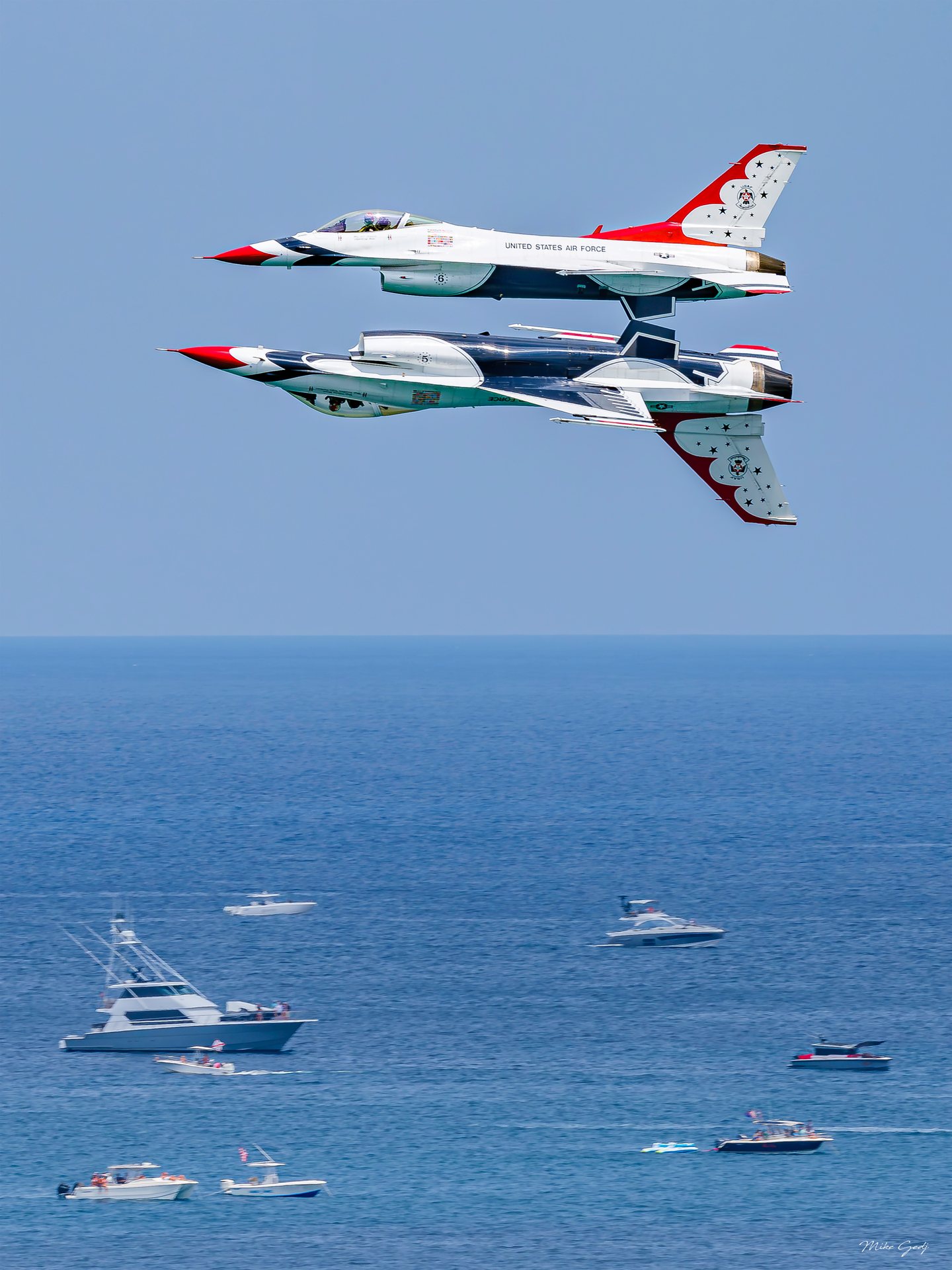 Two red, white and blue jets fly close together, one inverted, above a blue sea dotted with motorboats.