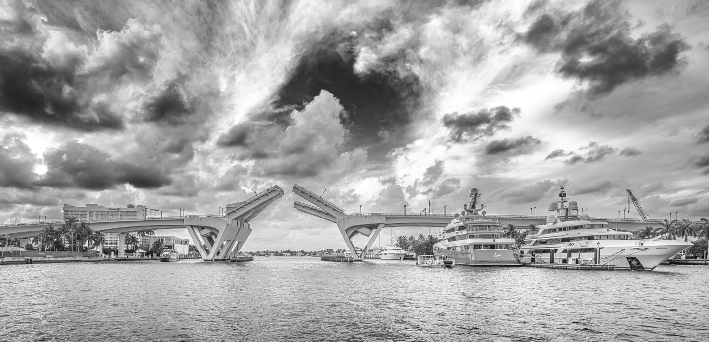 Black-and-white view of a raised drawbridge over rippled water, with large yachts, palms, and dramatic clouds.