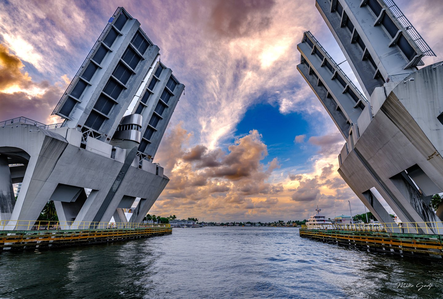 Raised drawbridge spans frame a waterway, yachts, and palm-lined shores beneath pink and gold clouds.
