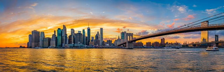 A suspension bridge crosses a high-rise waterfront beneath golden sunset clouds, with boats on the rippled river.