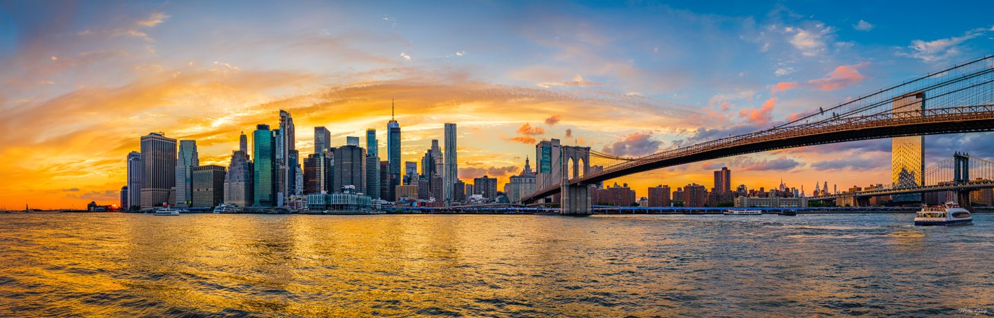 A suspension bridge crosses a high-rise waterfront beneath golden sunset clouds, with boats on the rippled river.