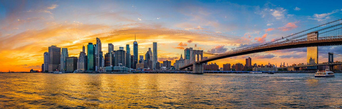 A suspension bridge crosses a high-rise waterfront beneath golden sunset clouds, with boats on the rippled river.