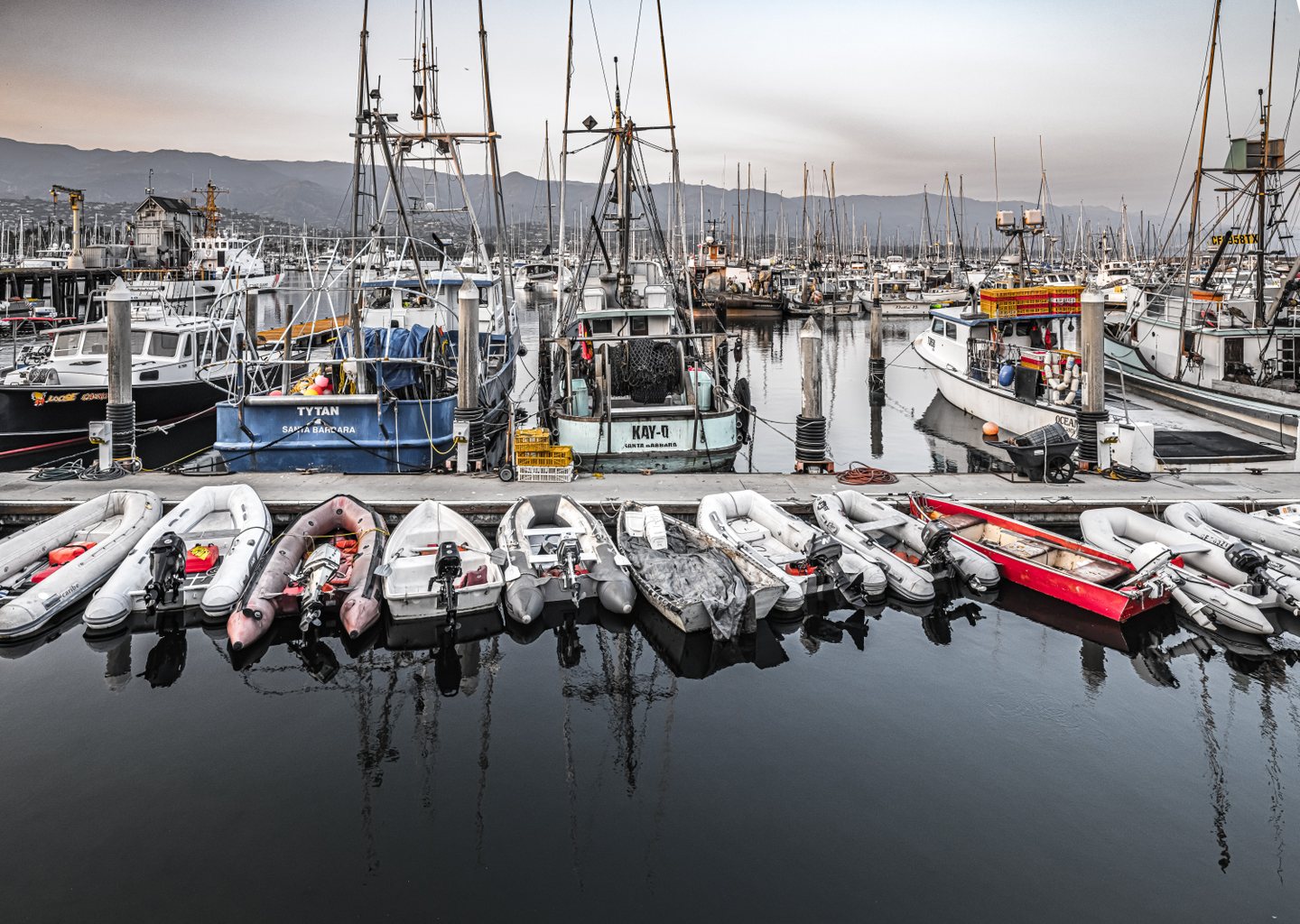 Dinghies crowd a dock before fishing boats and tall masts, with mountain ridges behind and reflections below.