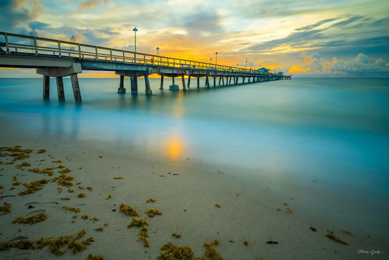 A lamp-lined pier crosses smooth turquoise water beneath golden clouds, with seaweed scattered across the sandy foreground.
