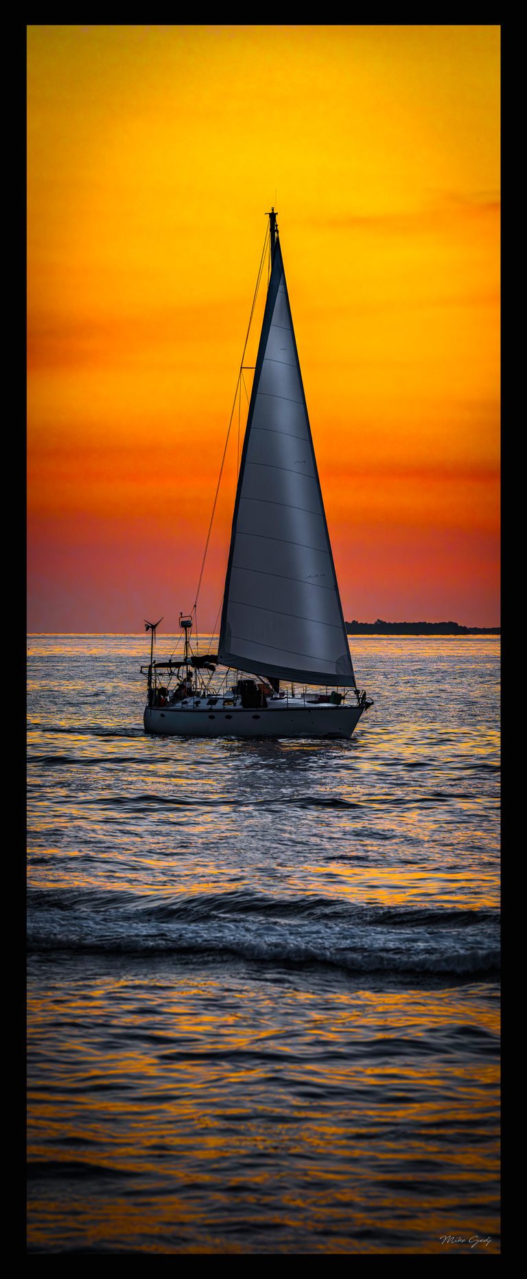 A sailboat with a tall gray sail crosses rippling water beneath a vivid orange sky and distant dark shoreline.