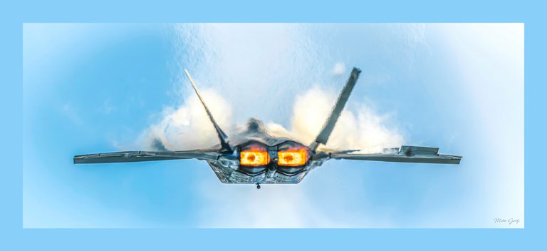 Rear view of a twin-engine fighter jet with glowing orange exhausts against a blue sky and white clouds.