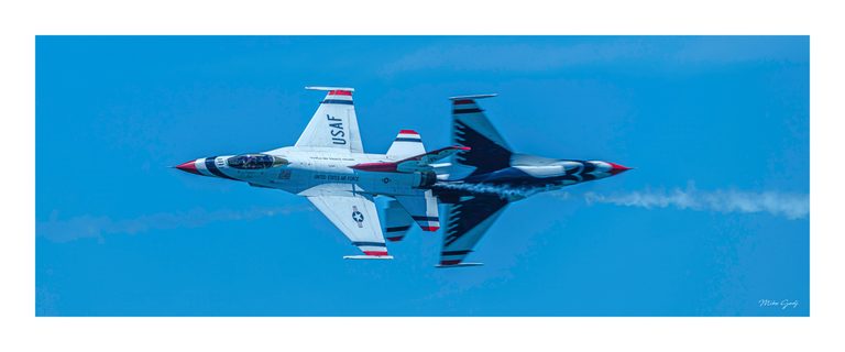 Two red, white, and blue U.S. Air Force jets pass in opposite directions against a blue sky, trailing white smoke.