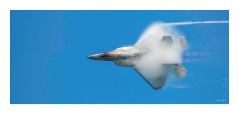 A gray fighter jet banks against a blue sky, its wings partly veiled in white vapor and its canopy gleaming amber.